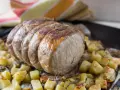 A tied roast pork loin sits on a bed of roasted potatoes and herbs on a baking sheet.