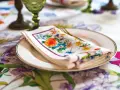 Tovagliolo fiorito piegato su un piatto bianco, con un bordo dorato. La tovaglia è decorata con un motivo floreale vivace. Il tovagliolo va a destra o sinistra?