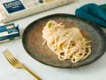 Un piatto di spaghetti cacio e pepe cremosi, guarniti con pepe nero macinato. Accanto, confezioni di pasta e burro, e una forchetta dorata.