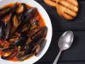 Un invitante piatto di zuppa di cozze in salsa rossa, accompagnato da crostini tostati e un cucchiaio d'argento.