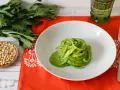 Piatto di spaghetti verdi con salsa cremosa, ispirato a ricette con foglie di sedano. Accanto, pinoli e un bicchiere verde.
