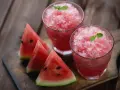 Due bicchieri di granita di anguria rinfrescante, guarniti con menta, accanto a fette di anguria su un tagliere di legno.