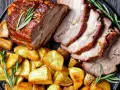 Lombata di maiale arrosto affettata, servita con patate dorate e rametti di rosmarino su un piatto scuro.
