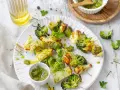 Spiedini di verdure grigliate, con broccoli, cavolfiore e cavolini di Bruxelles, conditi con pesto e serviti su un piatto bianco.