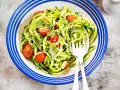 Deliziosi primi piatti con zucchine e pomodorini, conditi con pesto fresco e basilico, serviti in un piatto blu e bianco.