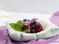 Due palline di gelato ai mirtilli, guarnite con mirtilli freschi e foglie di menta, servite in una ciotola bianca su un tovagliolo viola.