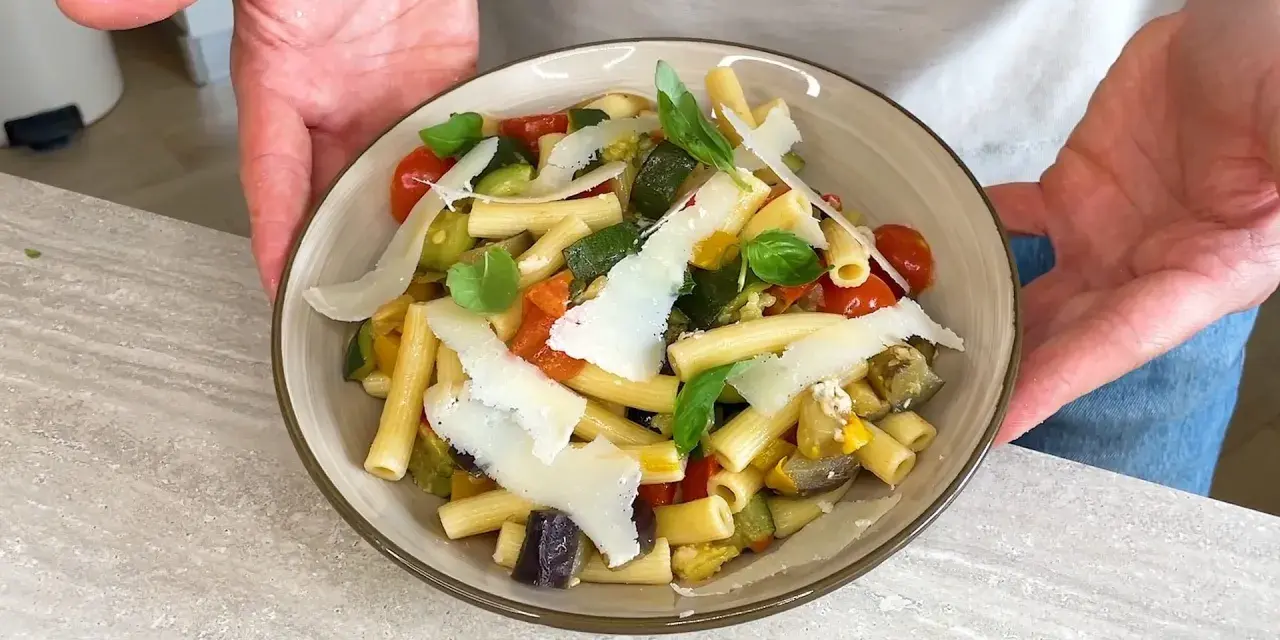 Un piatto di pasta fredda gourmet con verdure miste, pomodorini, basilico e scaglie di parmigiano.