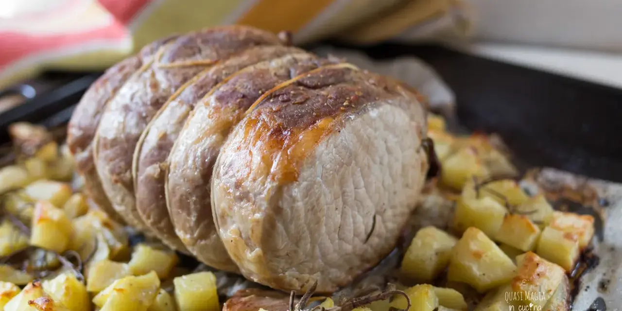 A tied roast pork loin sits on a bed of roasted potatoes and herbs on a baking sheet.