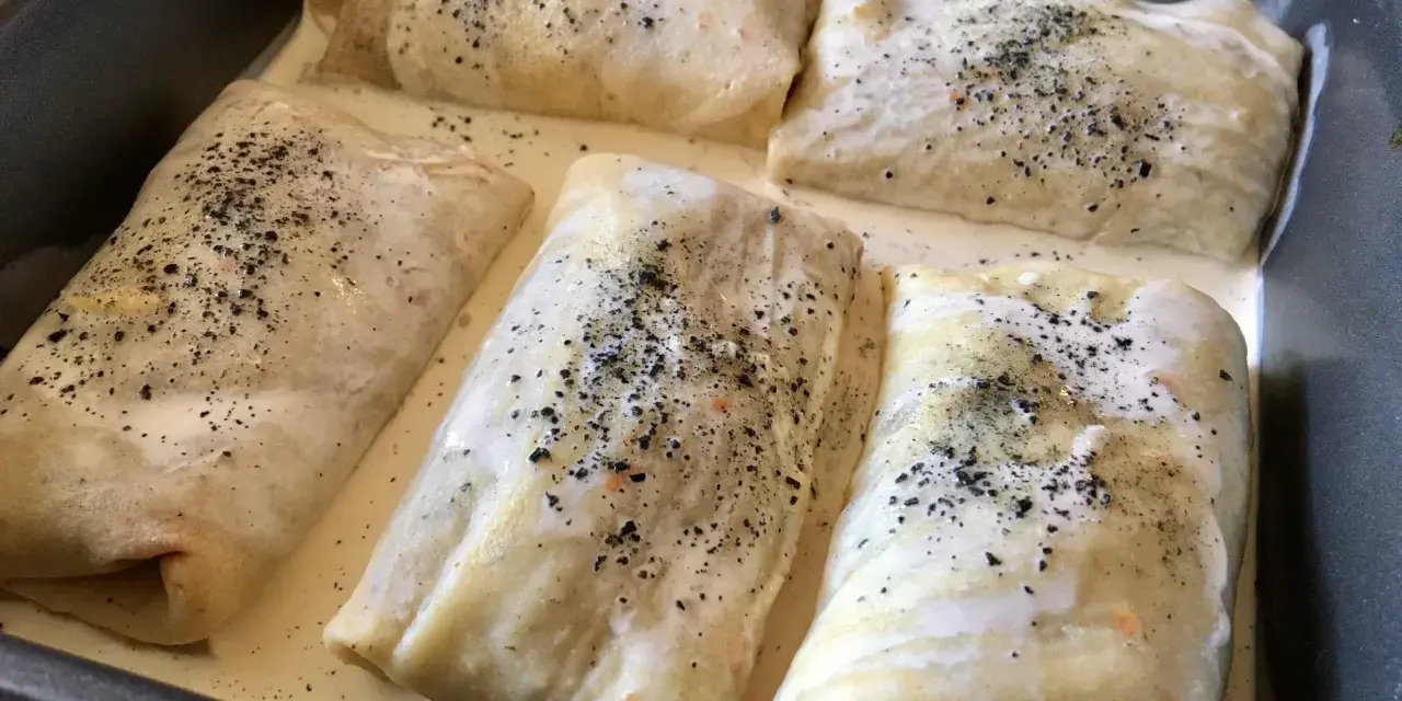 Crespelle farina di ceci ripiene, pronte per essere infornate, immerse in una cremosa besciamella e cosparse di pepe nero.