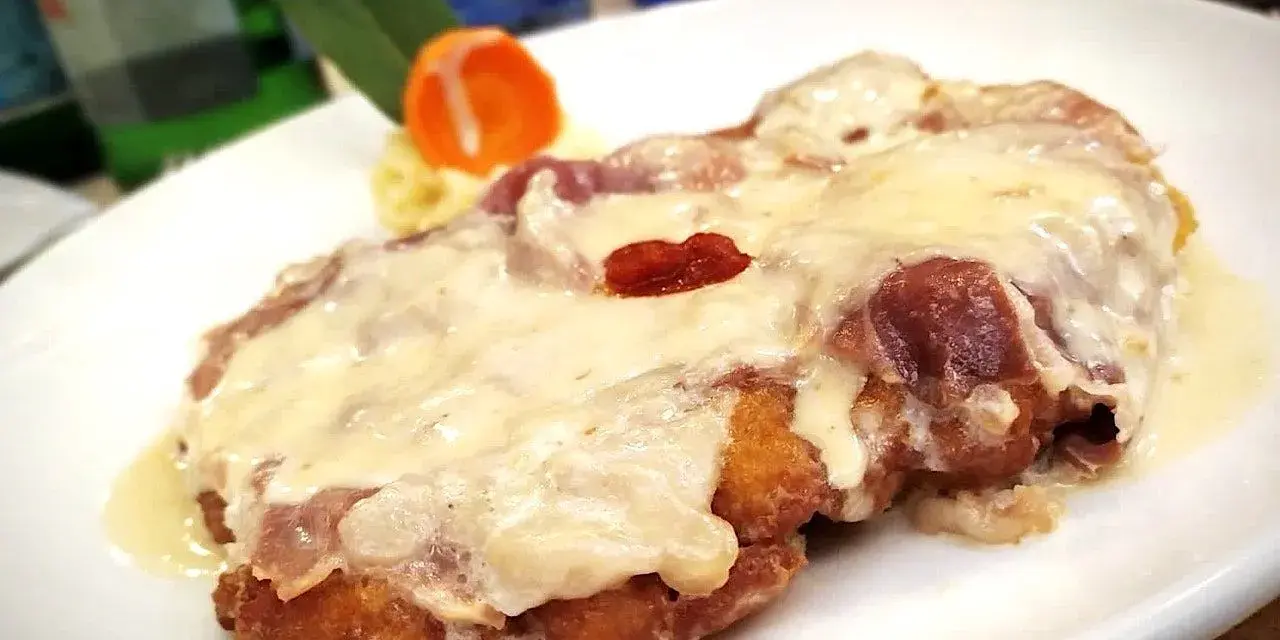 Cotoletta alla bolognese dorata e filante, con prosciutto e formaggio fuso, servita su un piatto bianco con contorno di purè e carota.