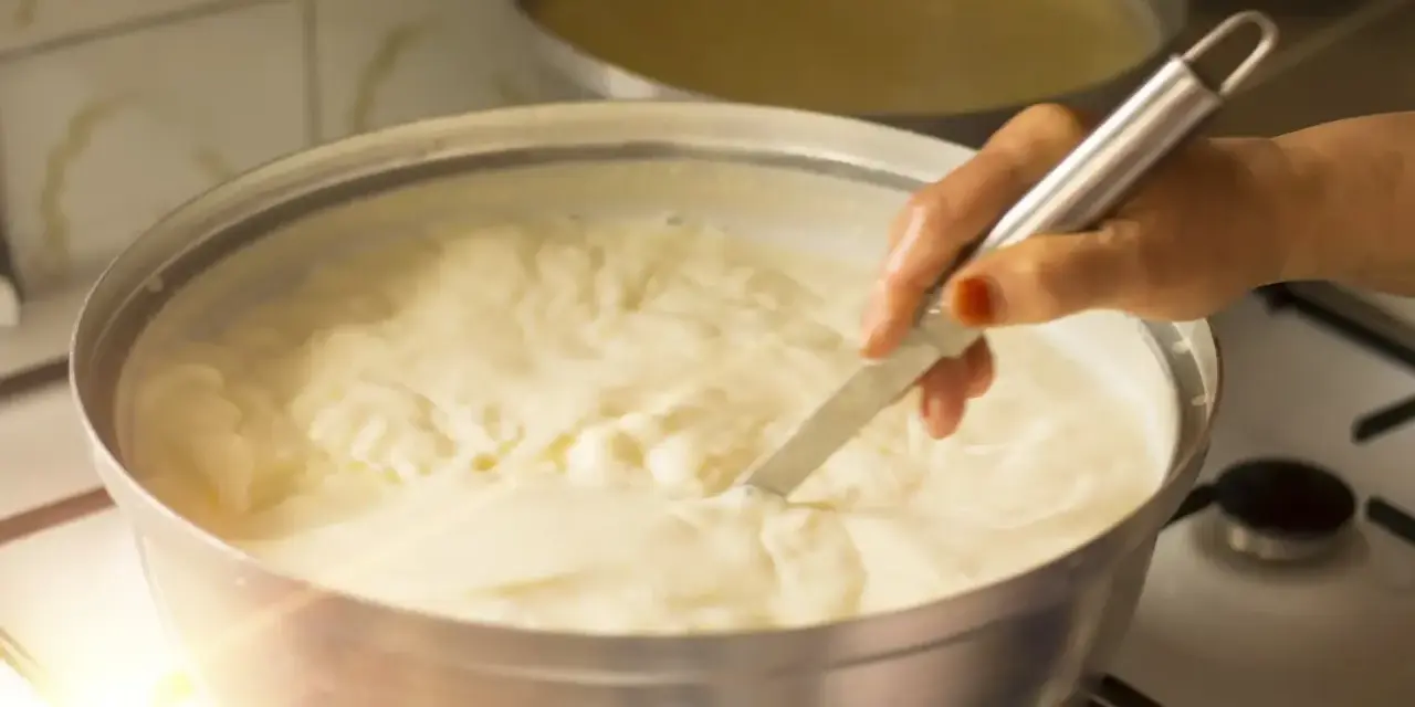 Una mano mescola un cremoso yogurt fatto in casa con fermenti in una ciotola d'acciaio su un fornello.