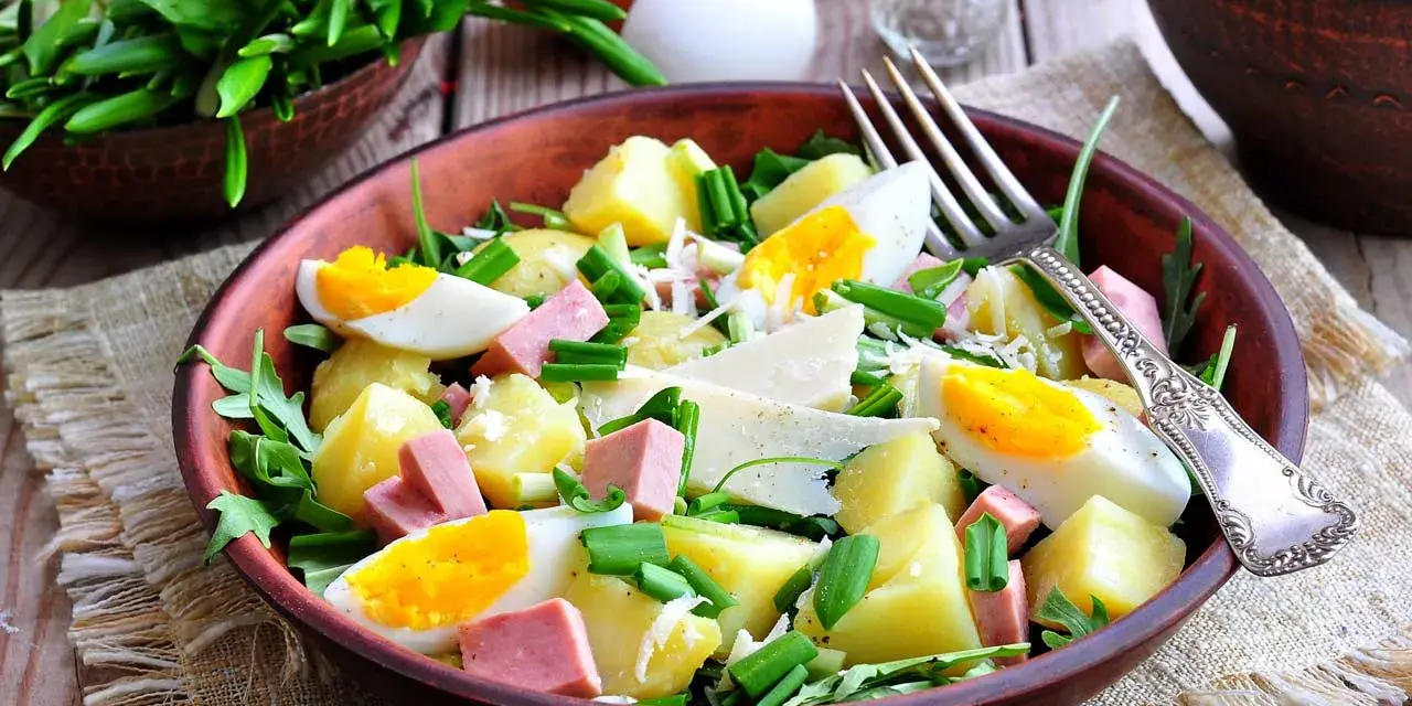 Deliziosa insalata di patate estiva con uova sode, salumi, formaggio e erbe fresche, servita in una ciotola di terracotta.