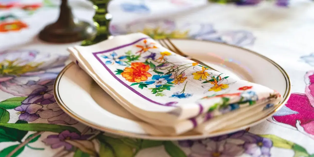 Tovagliolo fiorito piegato su un piatto bianco, con un bordo dorato. La tovaglia è decorata con un motivo floreale vivace. Il tovagliolo va a destra o sinistra?