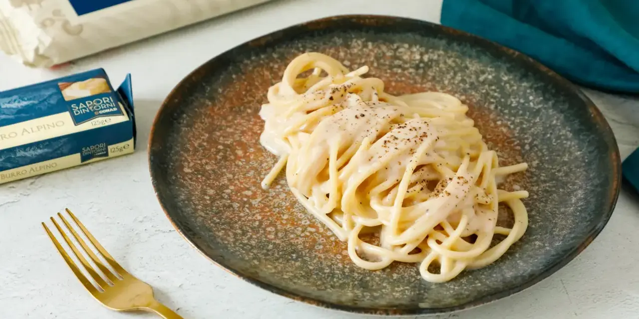 Un piatto di spaghetti cacio e pepe cremosi, guarniti con pepe nero macinato. Accanto, confezioni di pasta e burro, e una forchetta dorata.