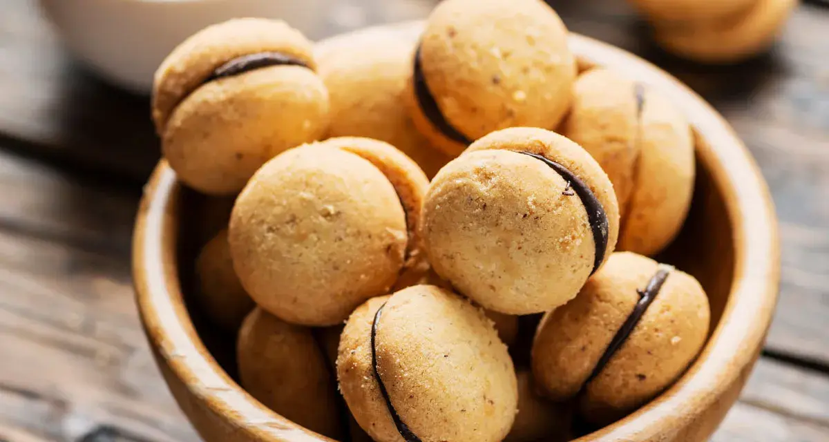 Baci di dama ricetta: deliziosi biscotti alle nocciole con ripieno di cioccolato, serviti in una ciotola di legno.
