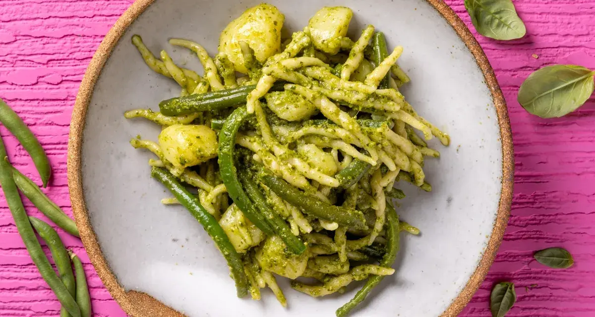 Un piatto invitante di trofie al pesto, con fagiolini e patate, su uno sfondo rosa vibrante.