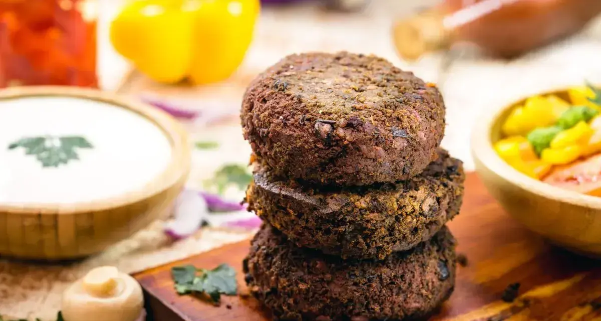 Tre gustosi hamburger di fagioli impilati su un tagliere di legno, con contorni colorati come salsa, peperoni e insalata.