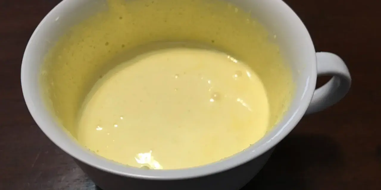 Una tazza bianca contiene un composto giallo pallido, un uovo sbattuto con zucchero, pronto per essere cucinato.
