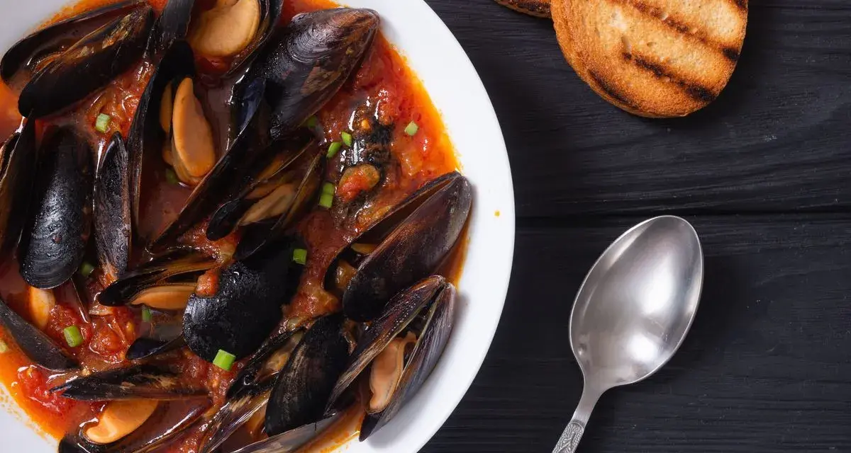 Un invitante piatto di zuppa di cozze in salsa rossa, accompagnato da crostini tostati e un cucchiaio d'argento.
