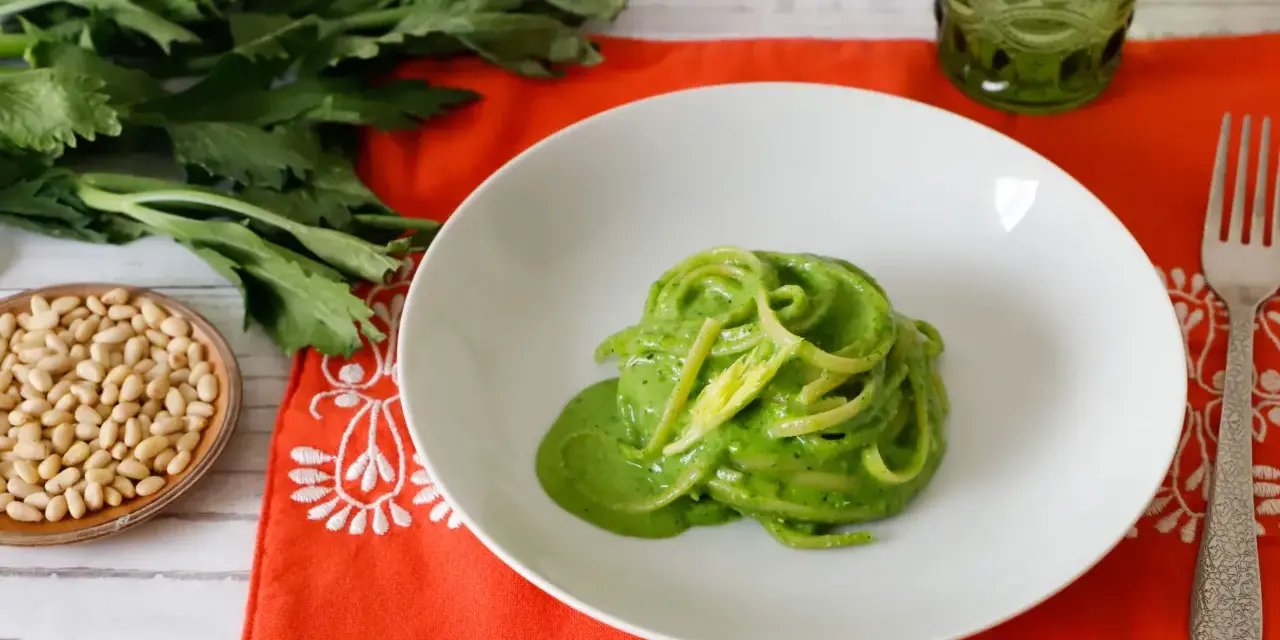 Piatto di spaghetti verdi con salsa cremosa, ispirato a ricette con foglie di sedano. Accanto, pinoli e un bicchiere verde.