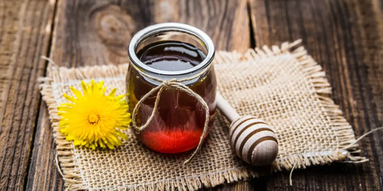 Un vasetto di miele ambrato, un fiore giallo e un cucchiaio di legno su tela grezza. La differenza tra miele e melata è un mistero da svelare.