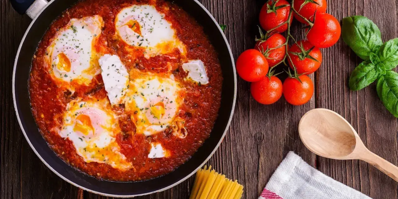 Uova al pomodoro in padella, con pomodorini freschi, basilico e un cucchiaio di legno.