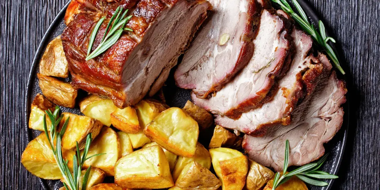 Lombata di maiale arrosto affettata, servita con patate dorate e rametti di rosmarino su un piatto scuro.
