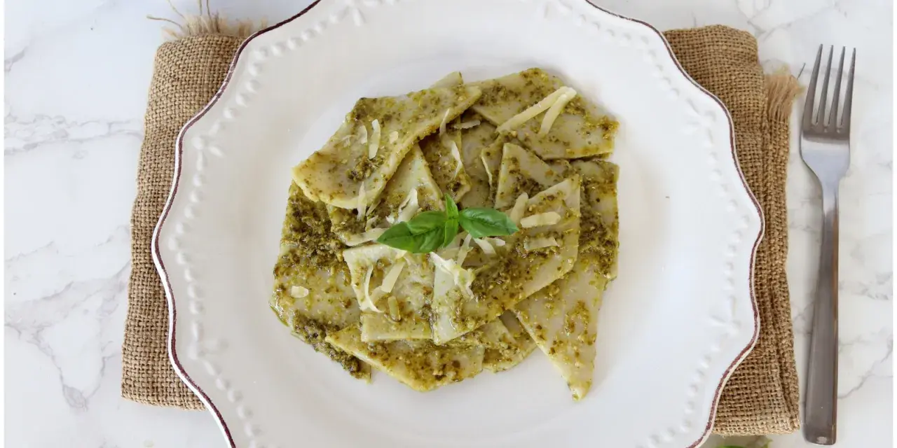 Testaroli al pesto, un piatto tradizionale ligure, servito su un piatto bianco con formaggio grattugiato e basilico fresco.