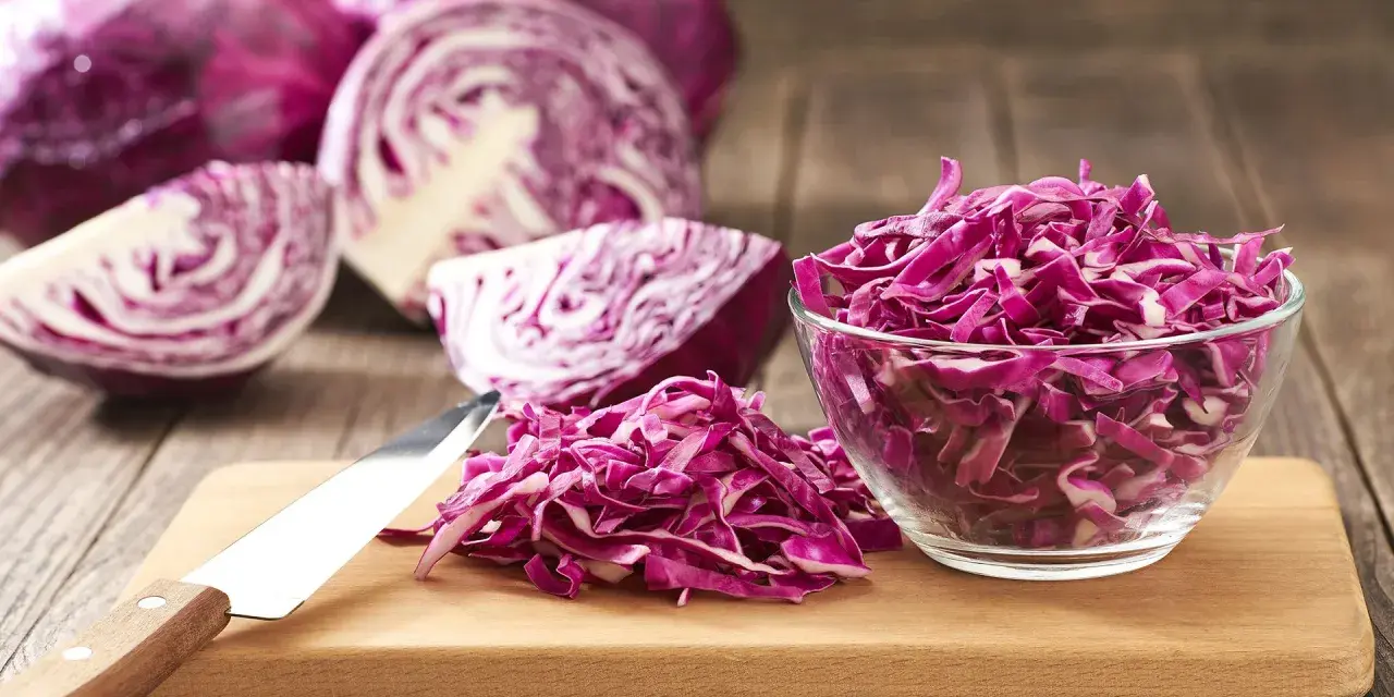 Cavolo cappuccio rosso affettato in una ciotola di vetro e su un tagliere, con cavoli interi e tagliati a spicchi sullo sfondo.