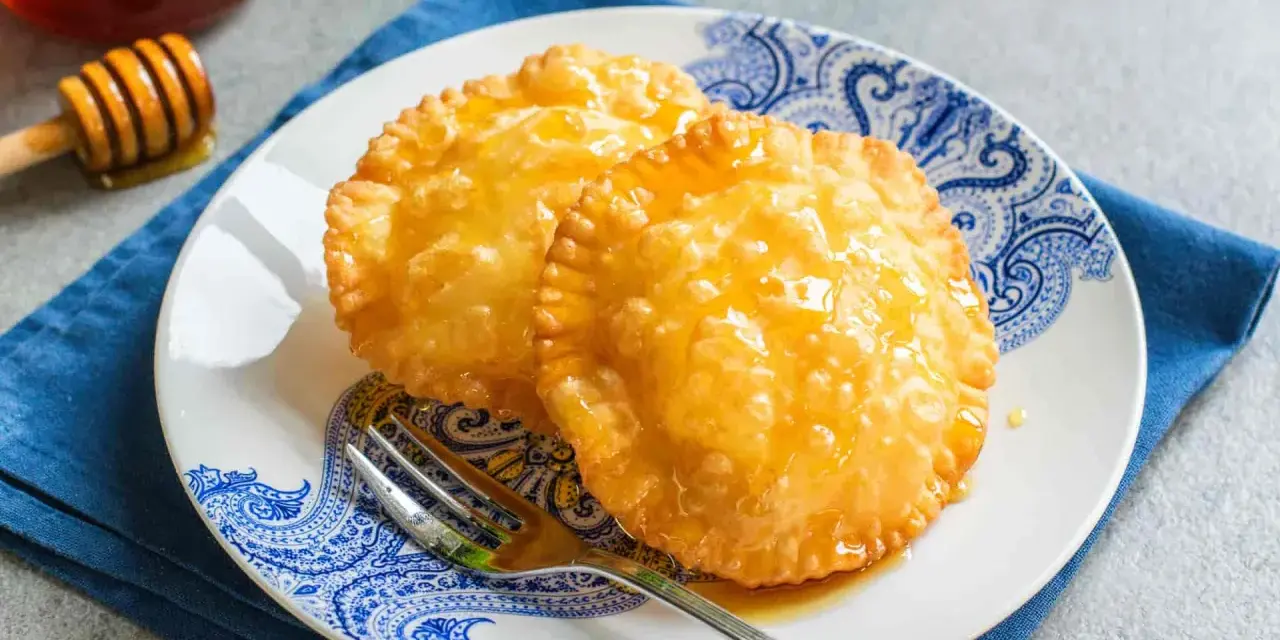 Due dolci sardi fritti, dorati e lucidi di miele, su un piatto blu e bianco con una forchetta.