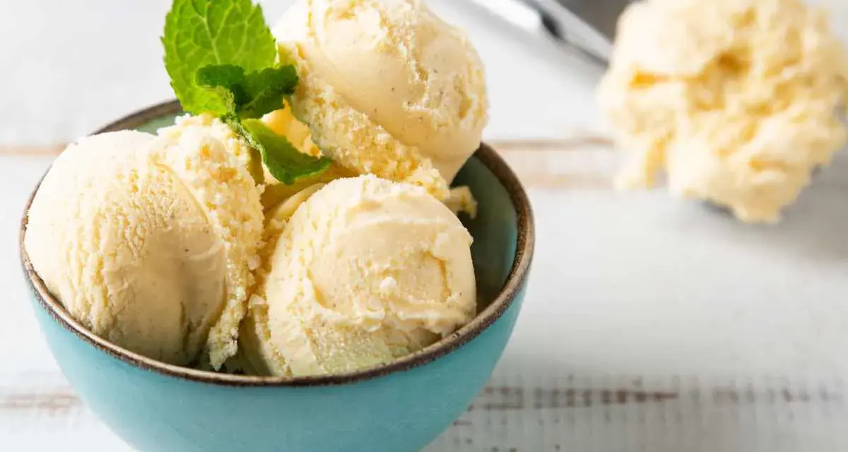 Coppa di cremoso gelato alla vaniglia, guarnita con foglioline di menta fresca. Sullo sfondo, un cucchiaio da gelato pieno.