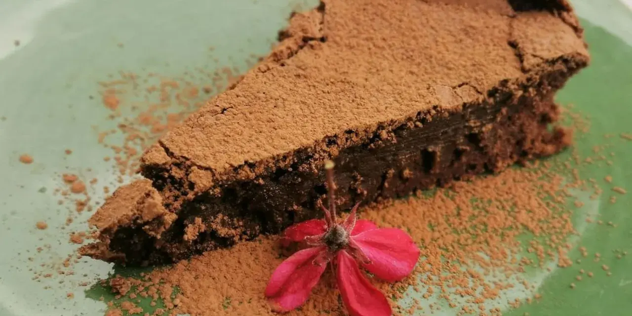 Una fetta di torta tenerina vegana, cosparsa di cacao, con un fiore rosso su un piatto verde.