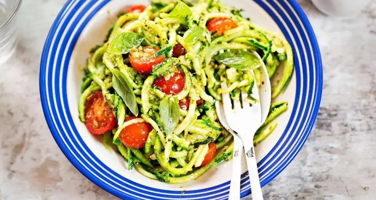 Deliziosi primi piatti con zucchine e pomodorini, conditi con pesto fresco e basilico, serviti in un piatto blu e bianco.