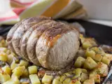 A tied roast pork loin sits on a bed of roasted potatoes and herbs on a baking sheet.