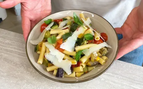 Un piatto di pasta fredda gourmet con verdure miste, pomodorini, basilico e scaglie di parmigiano.