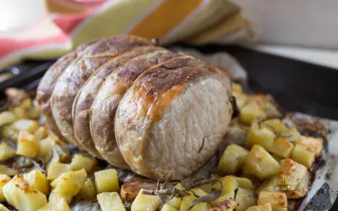 A tied roast pork loin sits on a bed of roasted potatoes and herbs on a baking sheet.