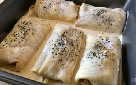 Crespelle farina di ceci ripiene, pronte per essere infornate, immerse in una cremosa besciamella e cosparse di pepe nero.