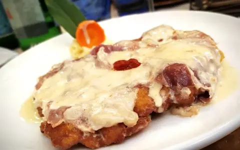 Cotoletta alla bolognese dorata e filante, con prosciutto e formaggio fuso, servita su un piatto bianco con contorno di purè e carota.