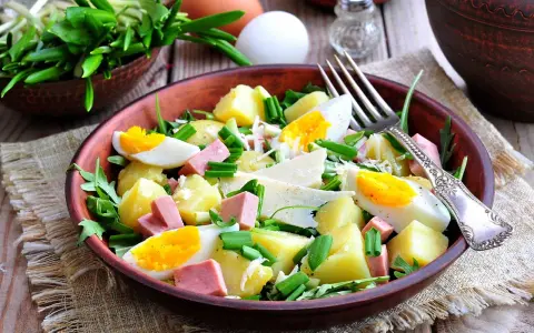 Deliziosa insalata di patate estiva con uova sode, salumi, formaggio e erbe fresche, servita in una ciotola di terracotta.