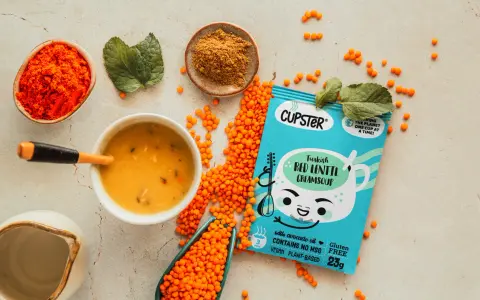 Una ciotola di zuppa lenticchie rosse cremosa, con lenticchie sparse, spezie e una confezione di zuppa istantanea.