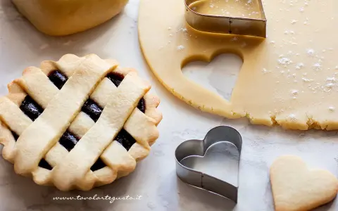 Preparazione di crostatine con pasta frolla classica. Stesa di impasto, formine a cuore e una crostatina già pronta.