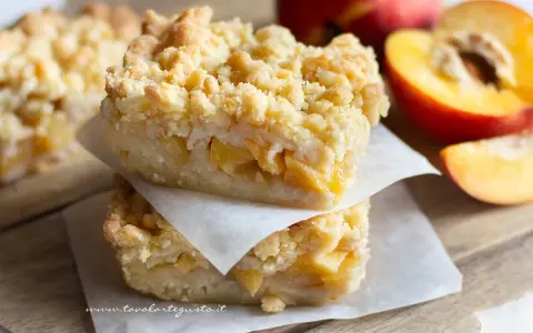 Sbriciolata pesche e amaretti: due fette impilate su carta forno, con pesche fresche sullo sfondo.