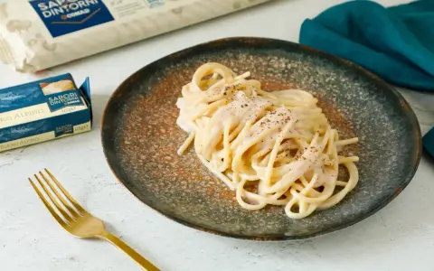 Un piatto di spaghetti cacio e pepe cremosi, guarniti con pepe nero macinato. Accanto, confezioni di pasta e burro, e una forchetta dorata.
