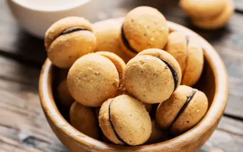 Baci di dama ricetta: deliziosi biscotti alle nocciole con ripieno di cioccolato, serviti in una ciotola di legno.