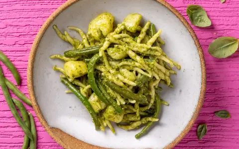 Un piatto invitante di trofie al pesto, con fagiolini e patate, su uno sfondo rosa vibrante.