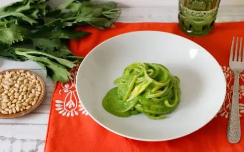 Piatto di spaghetti verdi con salsa cremosa, ispirato a ricette con foglie di sedano. Accanto, pinoli e un bicchiere verde.