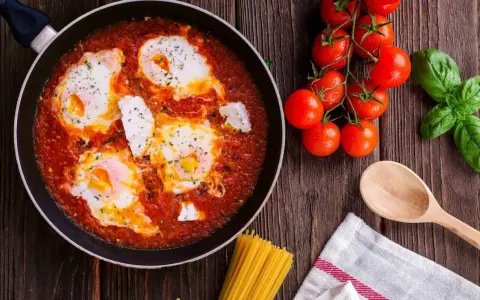 Uova al pomodoro in padella, con pomodorini freschi, basilico e un cucchiaio di legno.