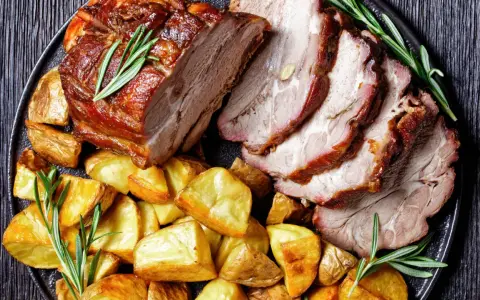 Lombata di maiale arrosto affettata, servita con patate dorate e rametti di rosmarino su un piatto scuro.