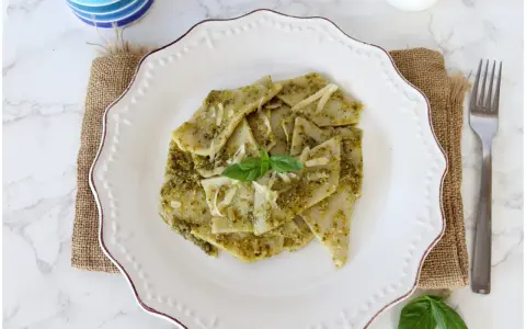 Testaroli al pesto, un piatto tradizionale ligure, servito su un piatto bianco con formaggio grattugiato e basilico fresco.