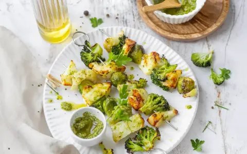 Spiedini di verdure grigliate, con broccoli, cavolfiore e cavolini di Bruxelles, conditi con pesto e serviti su un piatto bianco.